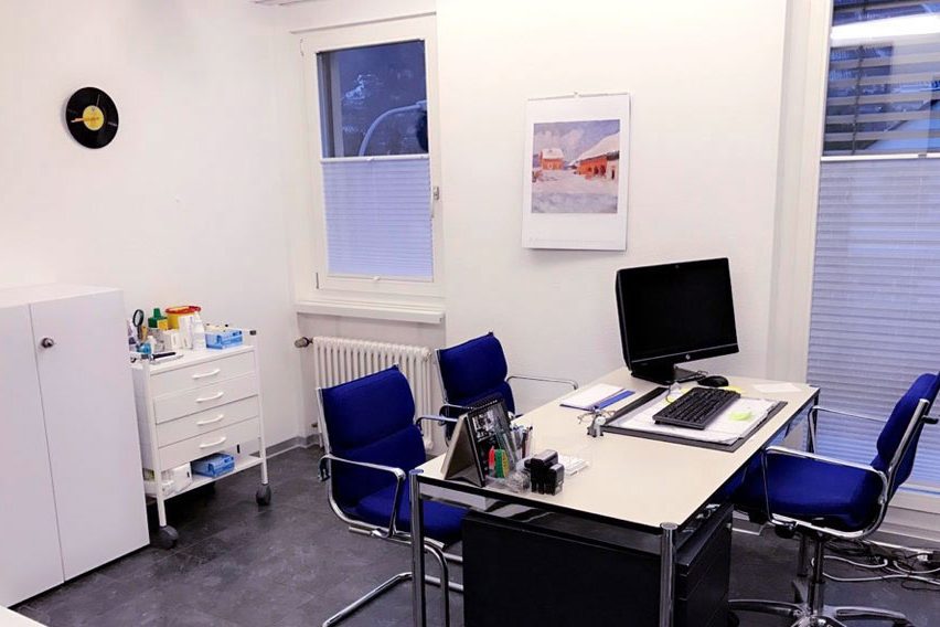 Sprechzimmer der Gemeinschaftspraxis Wettingen mit Schreibtisch, Bildschirm und Untersuchungsstuhl